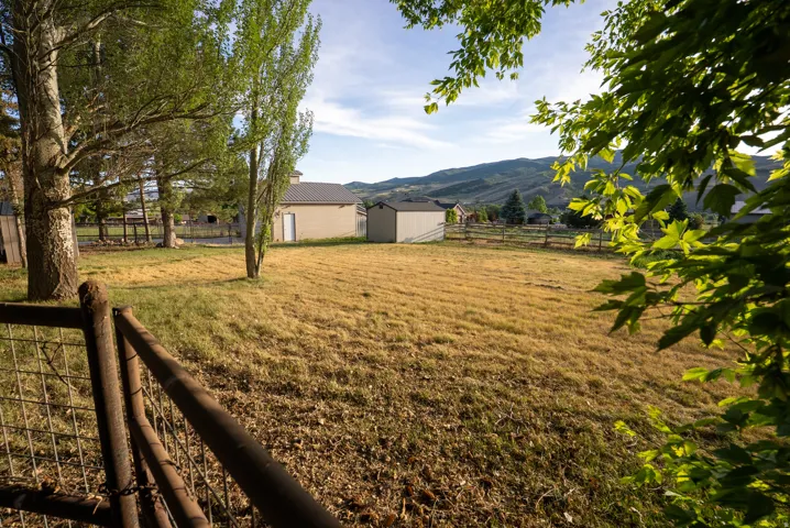 Expansive outdoor property featuring a rust-toned wire fence, mature trees, and a distant building with a dark-pitched roof
