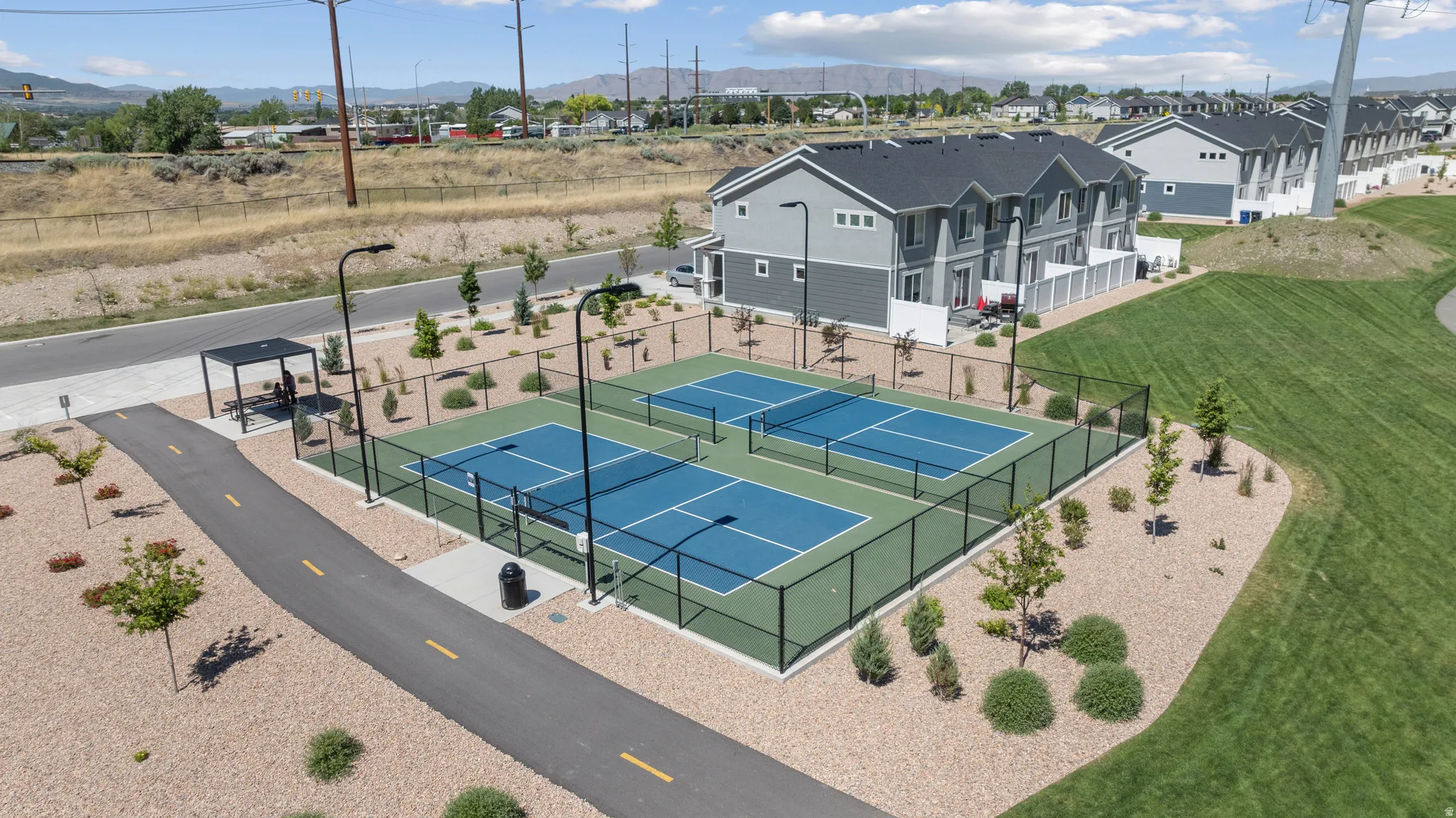 Community amenity area featuring two fenced pickleball courts with blue playing surfaces and green surrounds, a paved walking path, a shaded pergola, and manicured landscaping