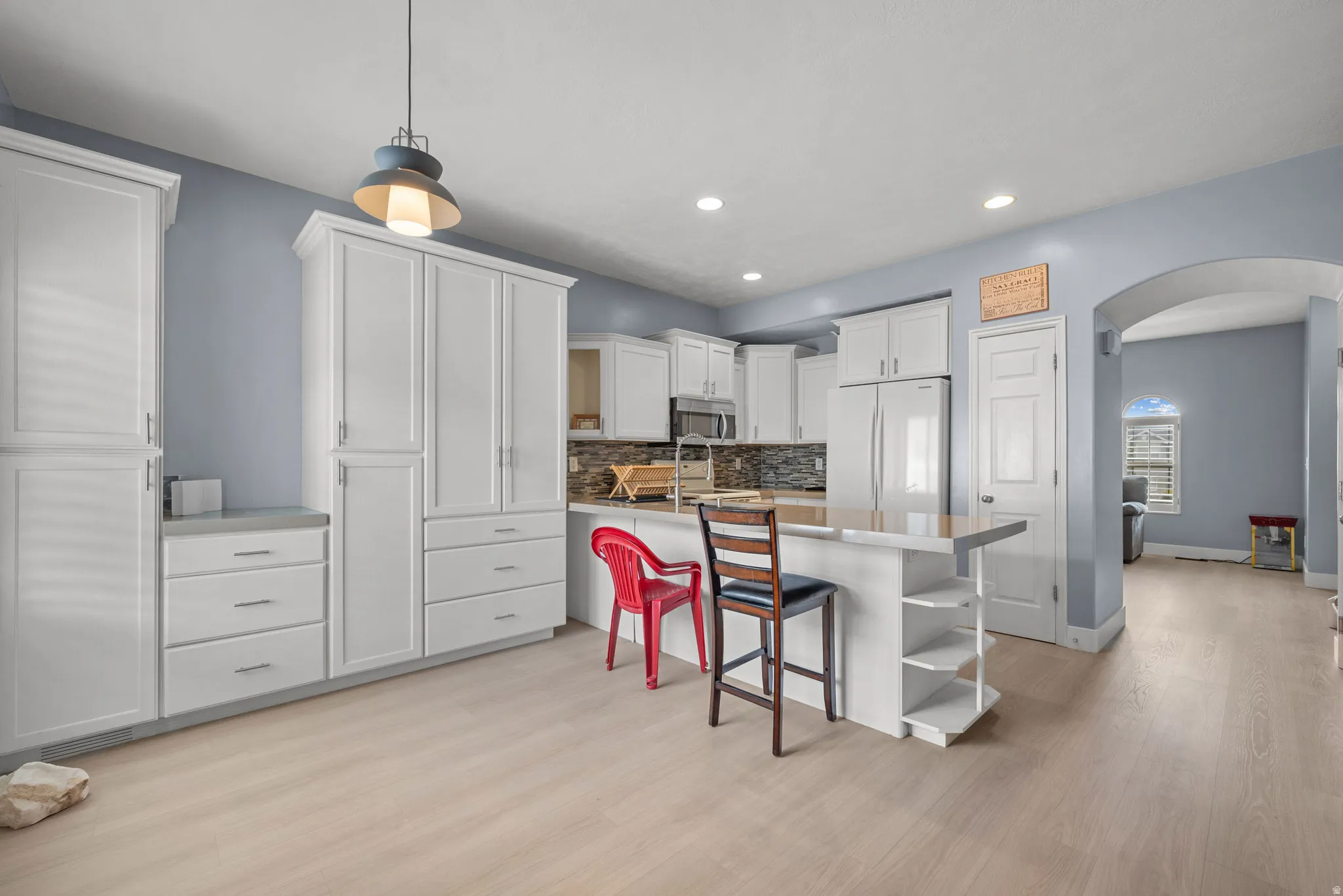 Bright kitchen featuring white shaker cabinetry, light wood-finish flooring, a central island with countertop seating, stainless steel refrigerator, and a mosaic tile backsplash