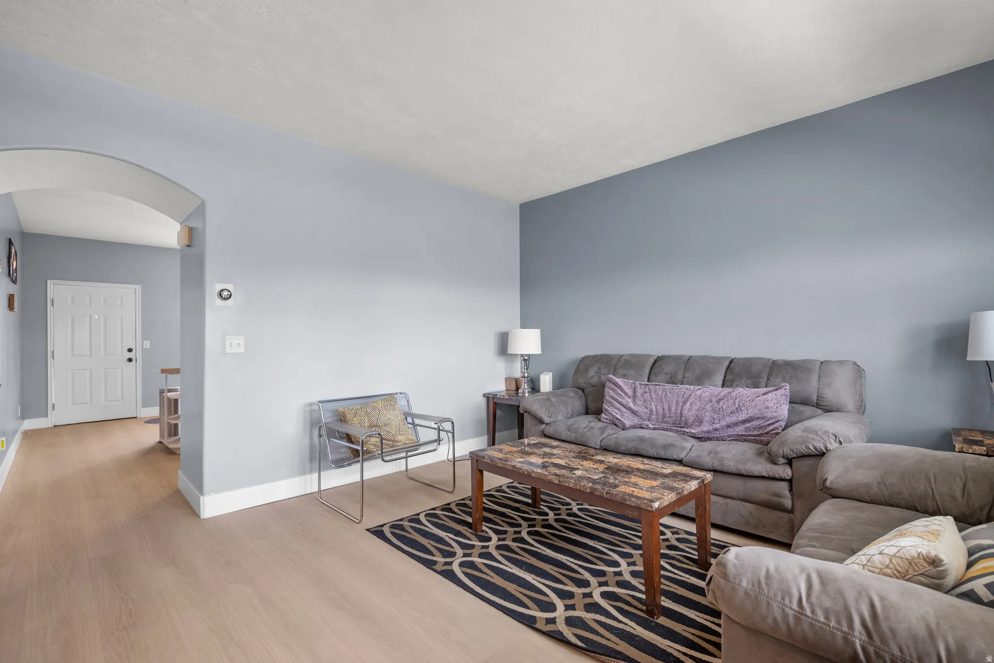 Living area featuring light wood-finish flooring, light blue wall paint, white baseboards, and a white paneled door visible through an arched doorway