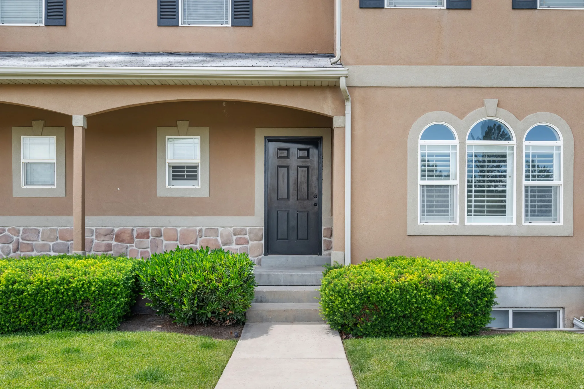 Entryway featuring a dark paneled door, arched window accents, stone foundation detailing, and manicured shrubbery