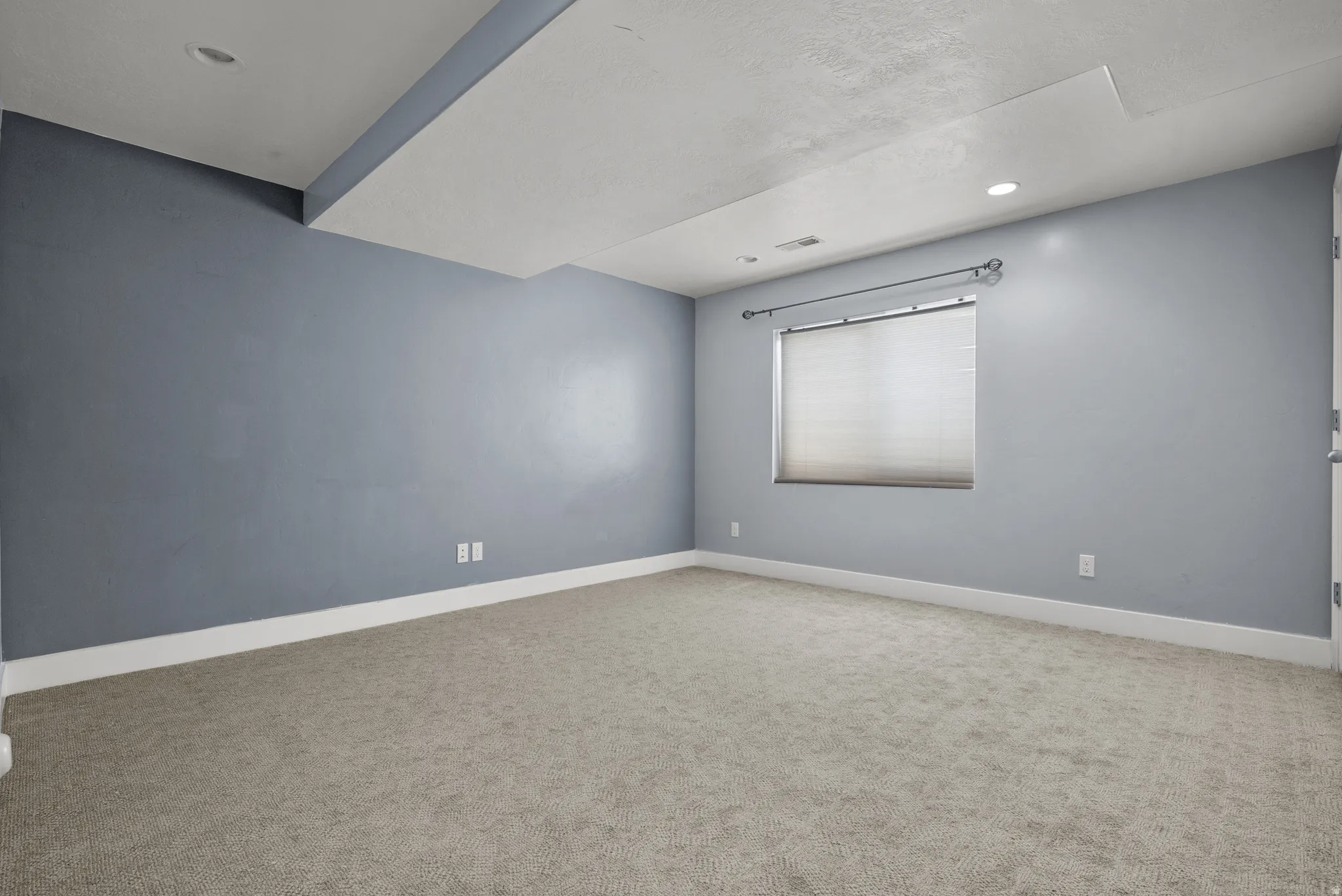 Spacious interior room featuring light gray carpeting, recessed ceiling lighting, and a window with blinds