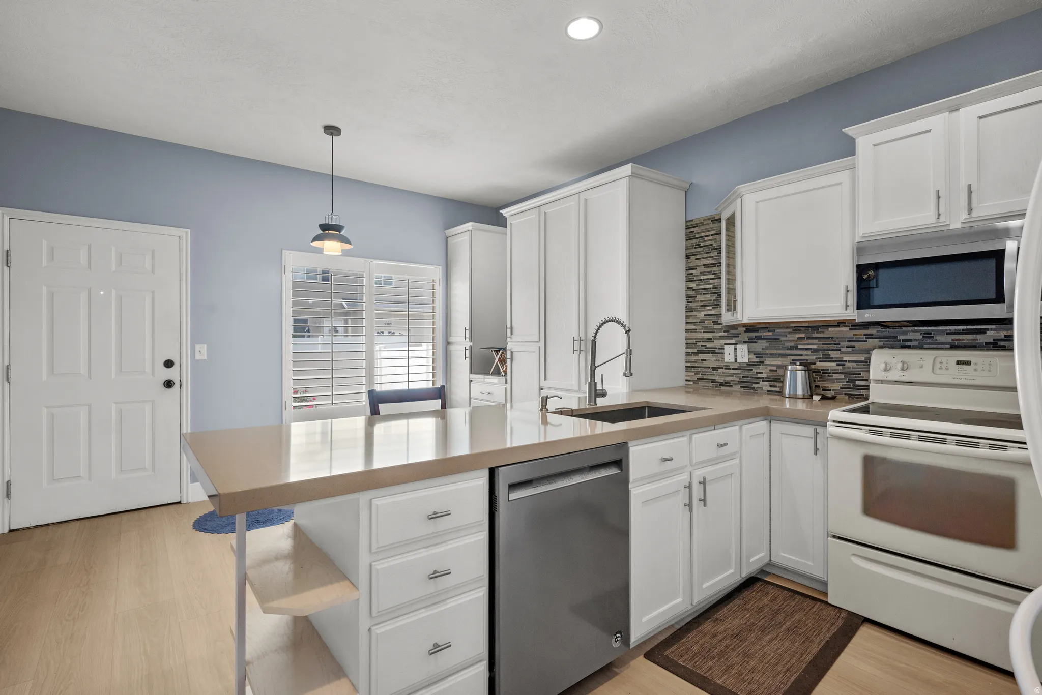 Kitchen featuring a central island with an integrated sink, light-toned countertops, white cabinetry with silver-tone hardware, a stainless steel dishwasher, and a tiled backsplash