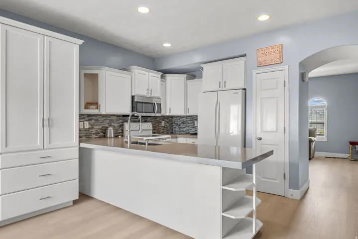 White shaker cabinetry, light-toned wood-finish flooring, kitchen island with integrated shelving, stainless steel appliances, and a mosaic tile backsplash