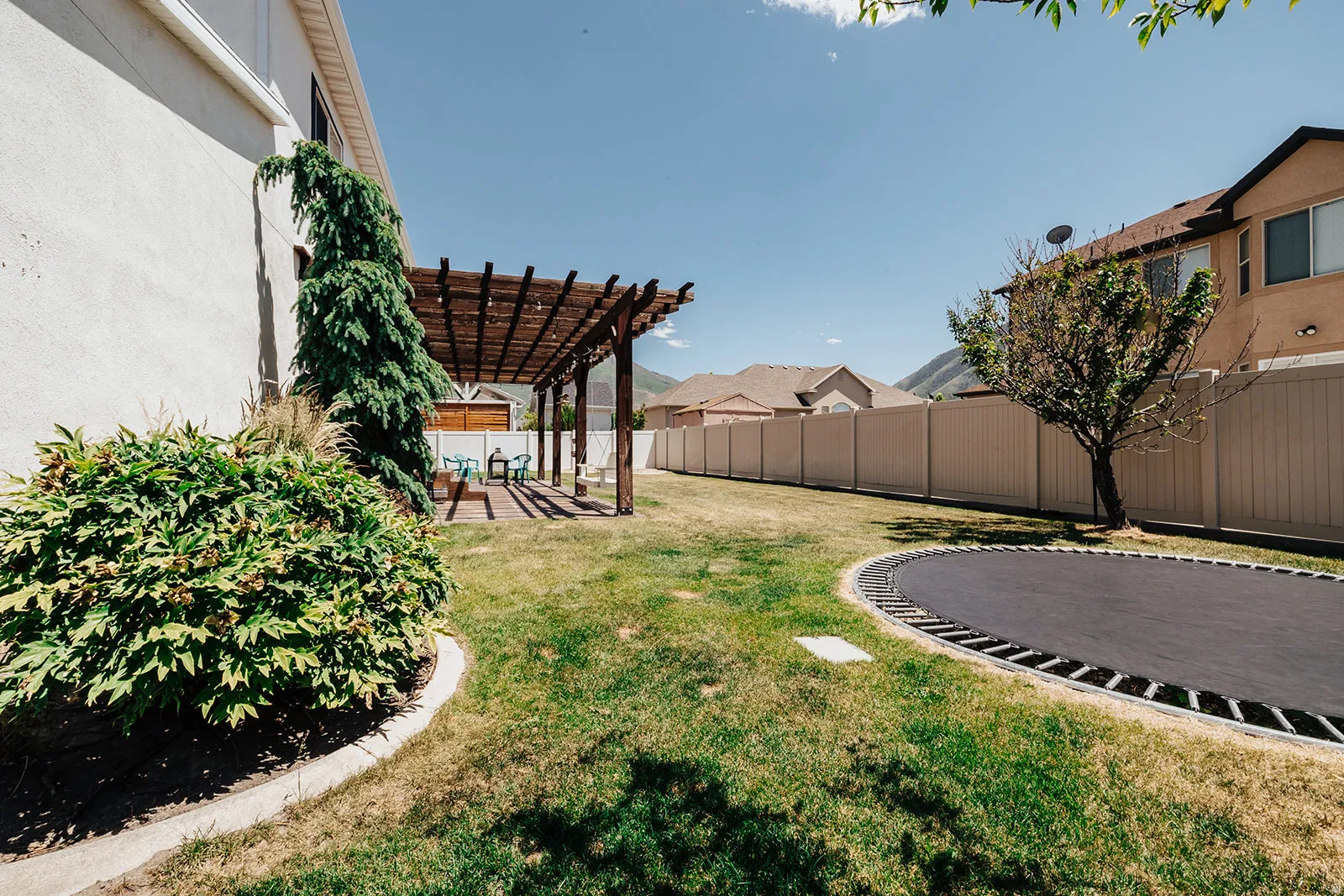 Spacious backyard featuring a wooden pergola, concrete patio, established landscaping, and a built-in ground trampoline