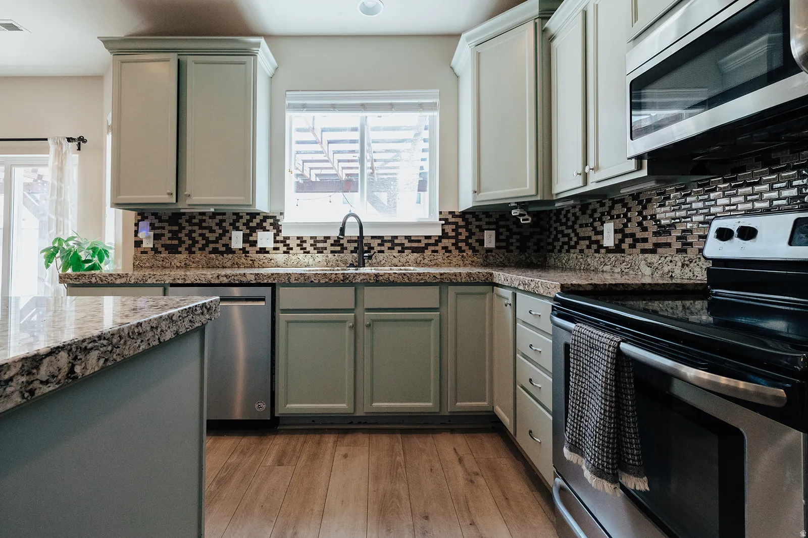 Kitchen featuring light green cabinetry, wood-finish flooring, stainless steel appliances, granite countertops, and a patterned tile backsplash