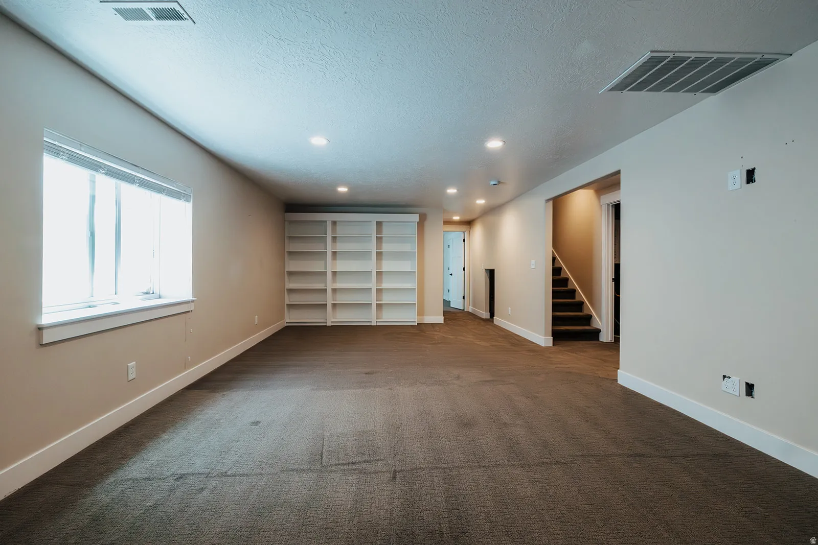 Spacious room featuring neutral carpeting, a large window with white trim, built-in shelving, recessed lighting, and a staircase with dark treads and white risers