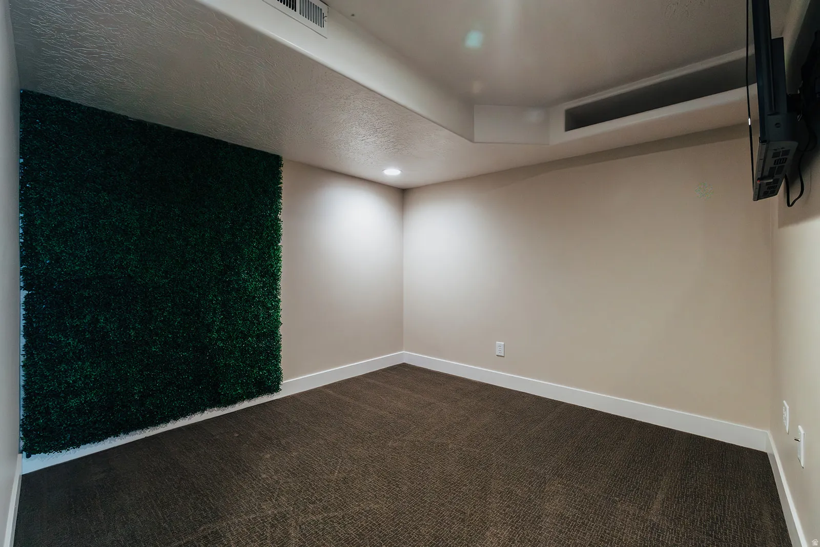 Accent wall featuring lush green artificial foliage, recessed ceiling lighting, textured ceiling, carpeted flooring, and crisp white baseboards