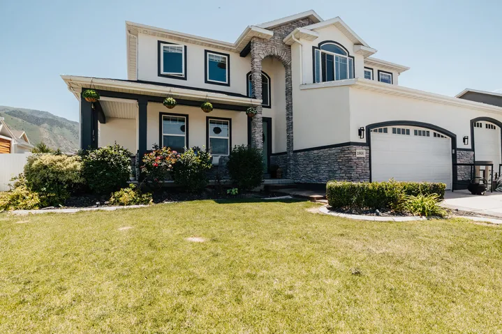 Two-story stucco and stone-clad residence featuring a covered front porch with dark columns, two arched garage doors, a stone archway entryway, and a verdant front lawn