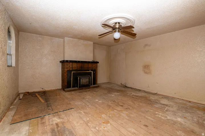 Living area featuring a brick fireplace with a black insert, wood-finish flooring, a ceiling fan with light fixture, textured walls, and an arched window