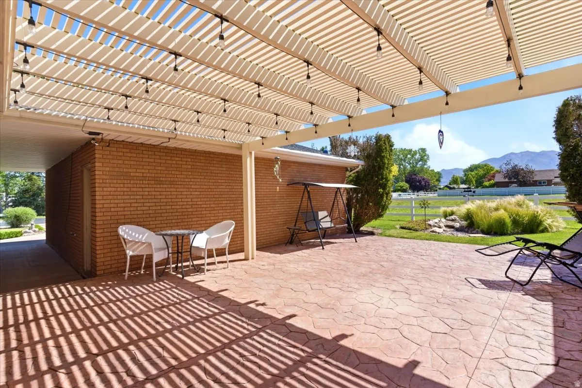 Expansive stamped concrete patio featuring a louvered patio cover with integrated string lights, brick exterior, and a manicured lawn with mountain backdrop