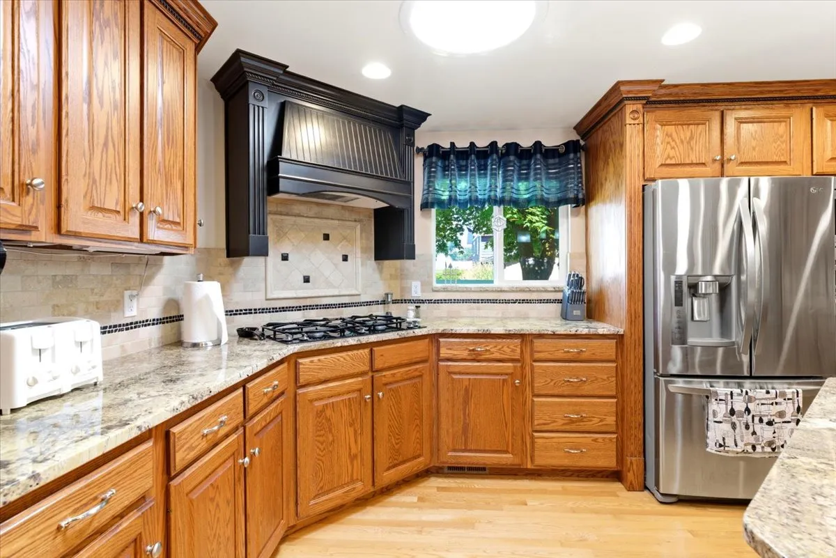 Kitchen featuring wood cabinetry, light-toned countertops, tile backsplash, stainless steel refrigerator, and a built-in gas cooktop with a decorative range hood