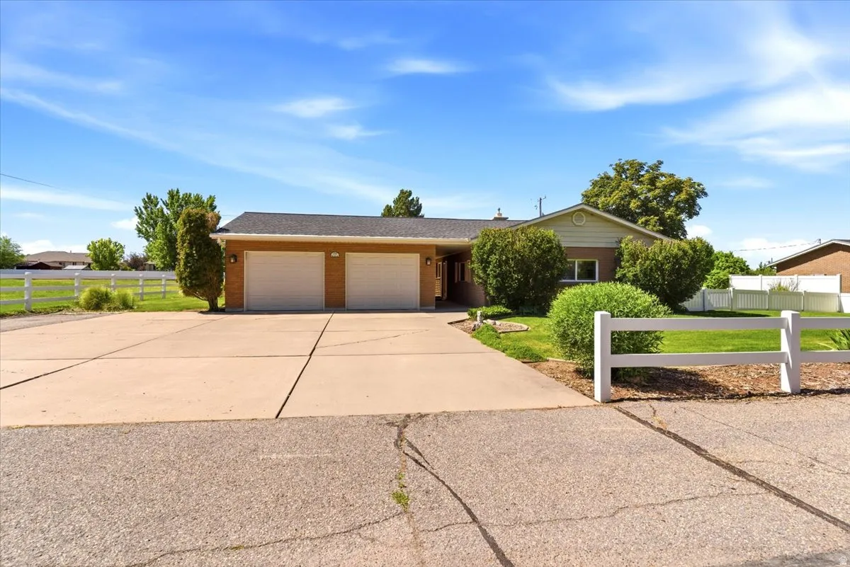 Brick exterior residence featuring a two-car garage, concrete driveway, white split-rail fence, mature landscaping, and a pitched roof