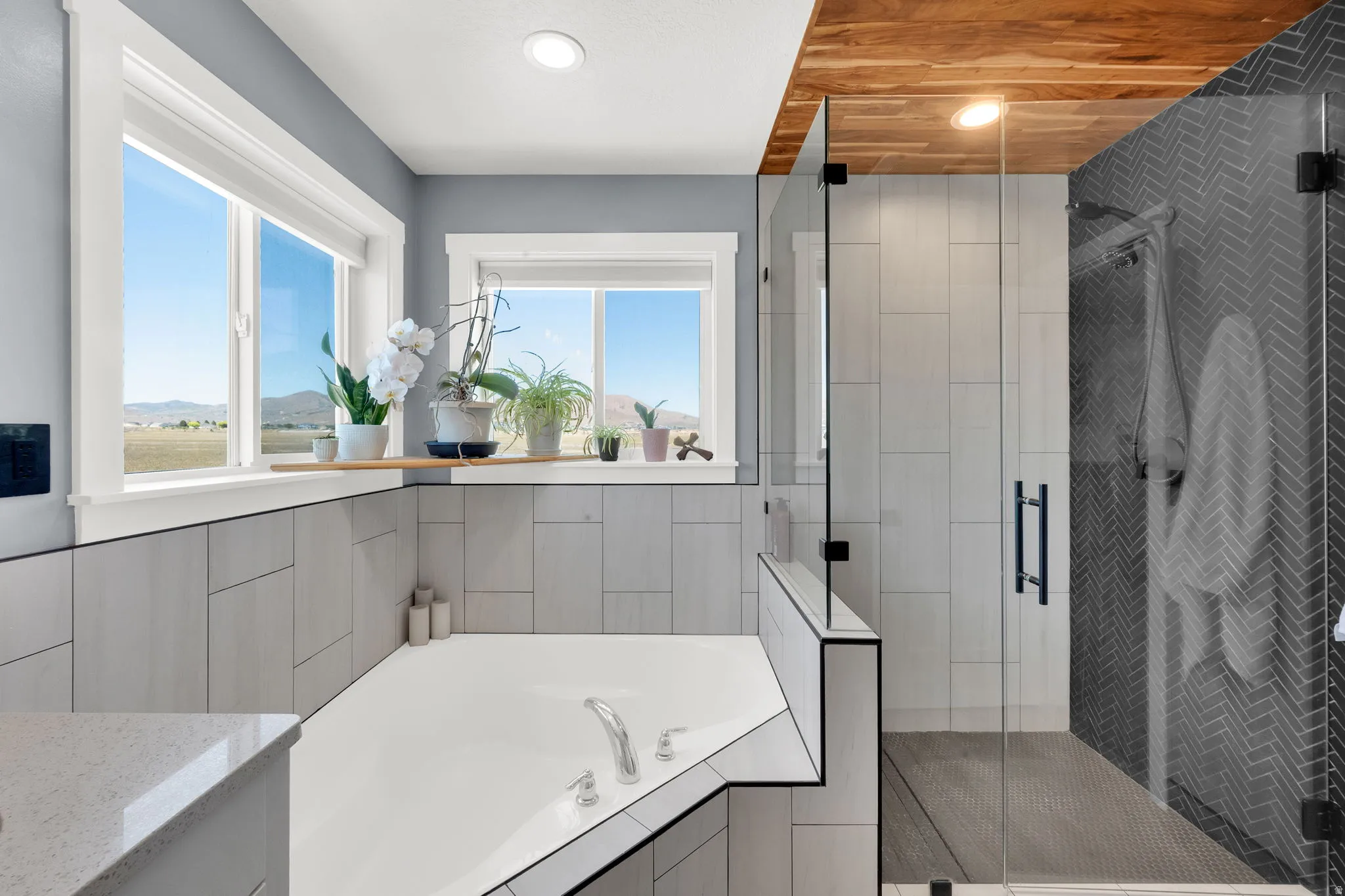 Freestanding soaking tub with integrated deck, surrounded by large windows, glass-enclosed shower with herringbone tile, and wood-paneled ceiling accent