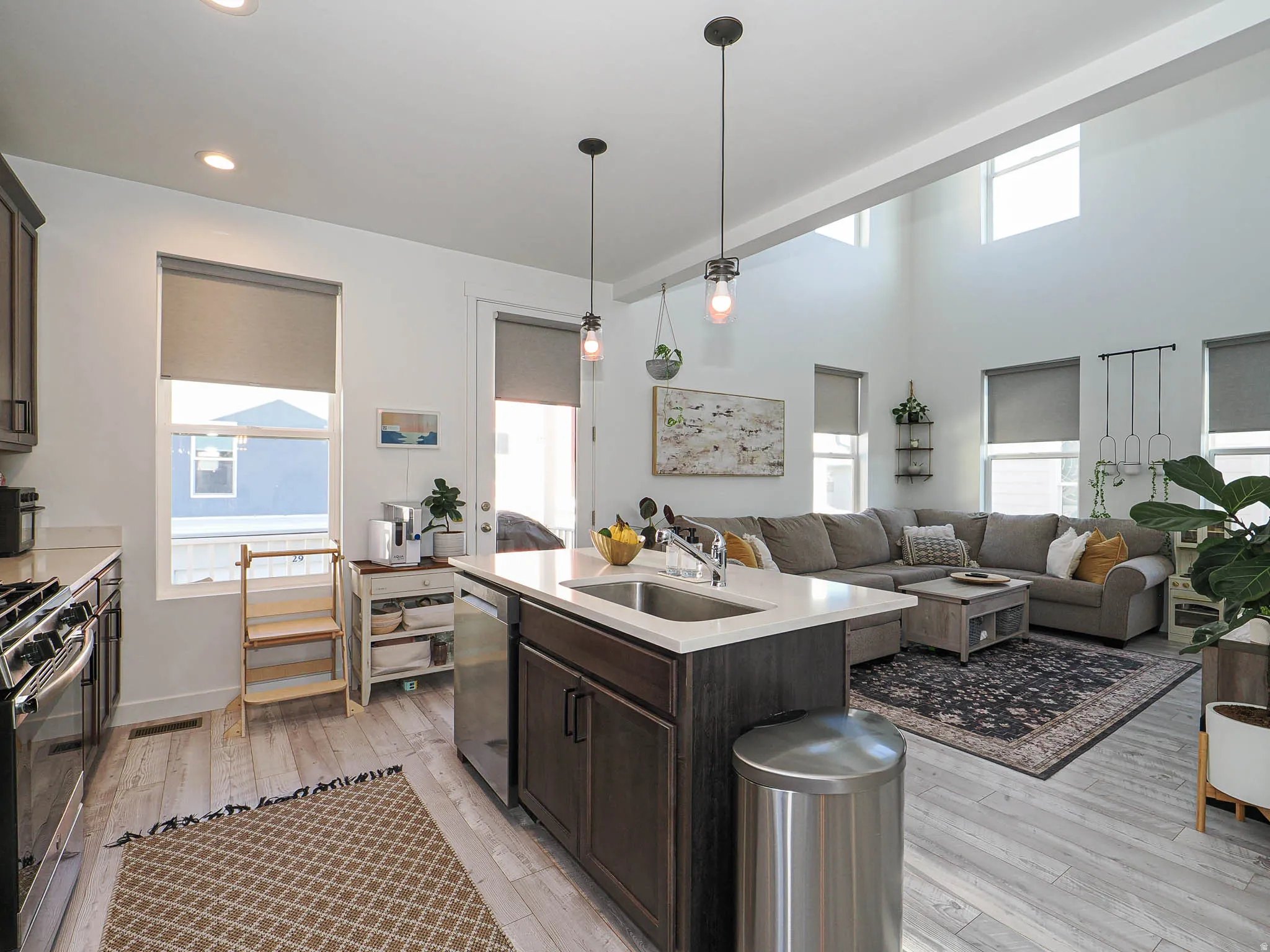 Open-concept living space featuring a kitchen island with sink, stainless steel appliances, wood-finish flooring, high ceilings, and multiple windows with roller shades