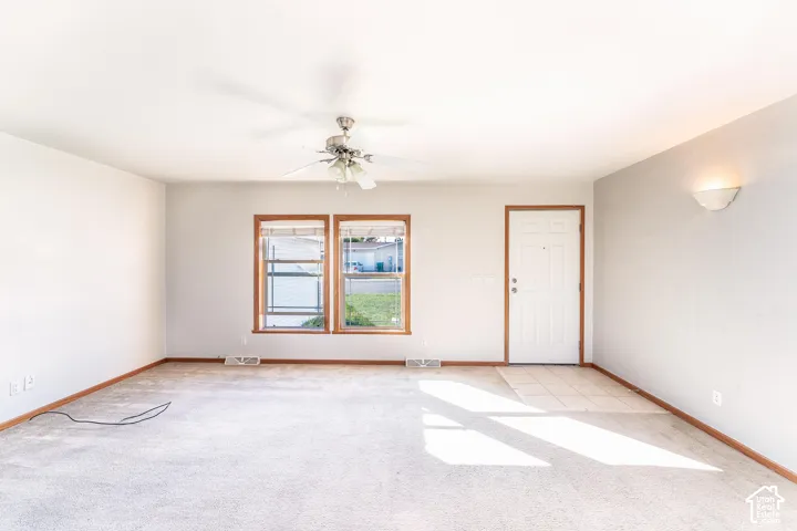 Spacious interior featuring neutral-toned carpeting, a ceiling fan with integrated lighting, dual-pane windows with wood-finish trim, and a tile-floored entryway