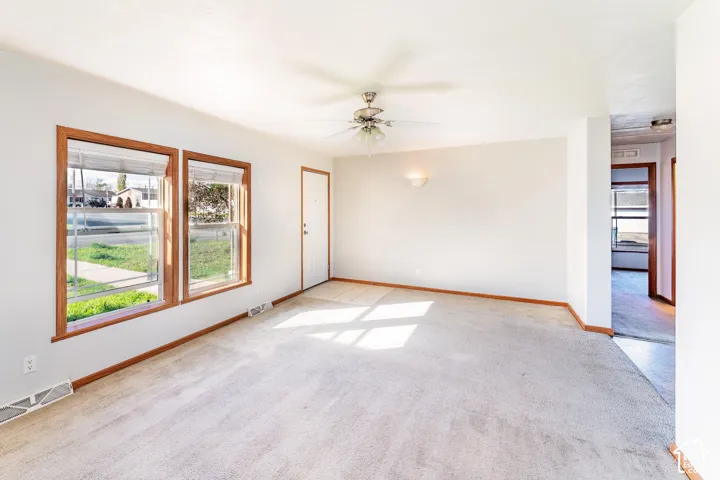 Living area featuring dual windows with wood-toned frames, neutral wall-to-wall carpeting, a ceiling fan with integrated lighting, and a wall-mounted sconce