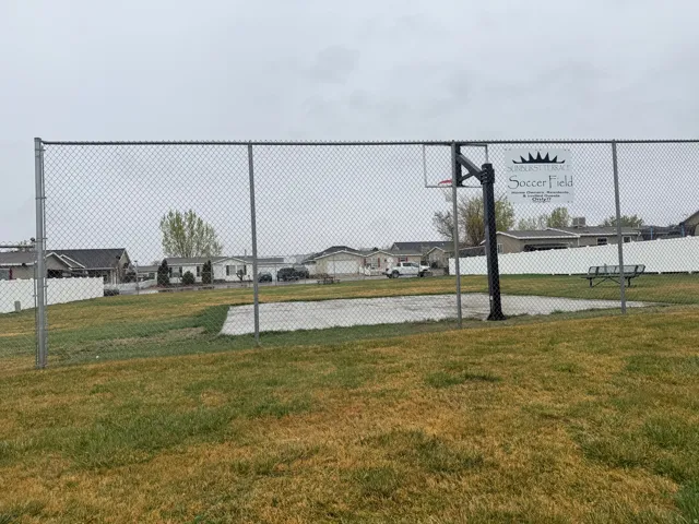 Chain-link fenced recreation area featuring a basketball hoop, concrete court, and a park bench