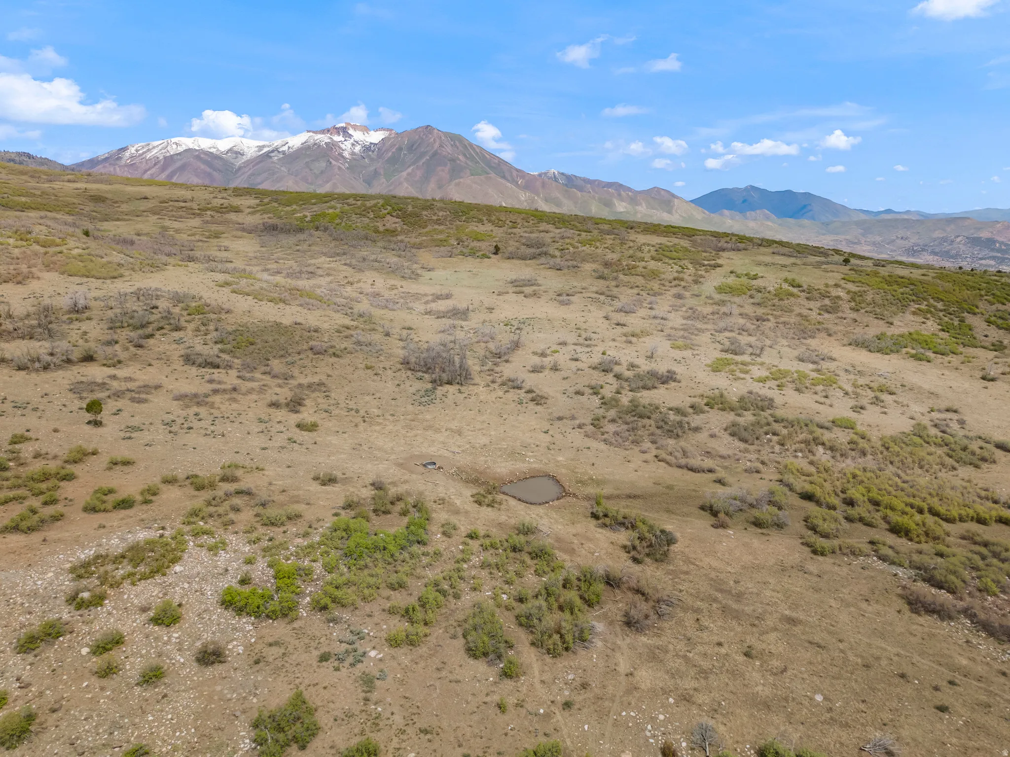 Expansive undeveloped acreage featuring varied terrain, a small natural pond, and distant snow-capped mountain peaks under a clear blue sky