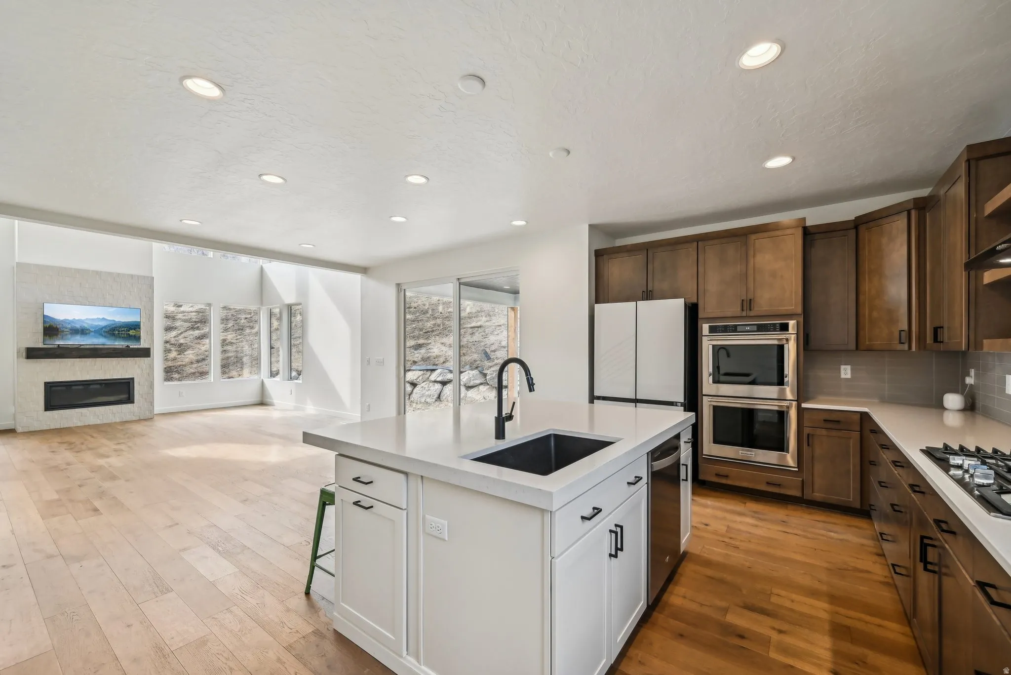 Open-concept kitchen and living area featuring a large center island with an integrated sink, light wood-finish flooring, recessed lighting, and a modern fireplace with a textured stone surround