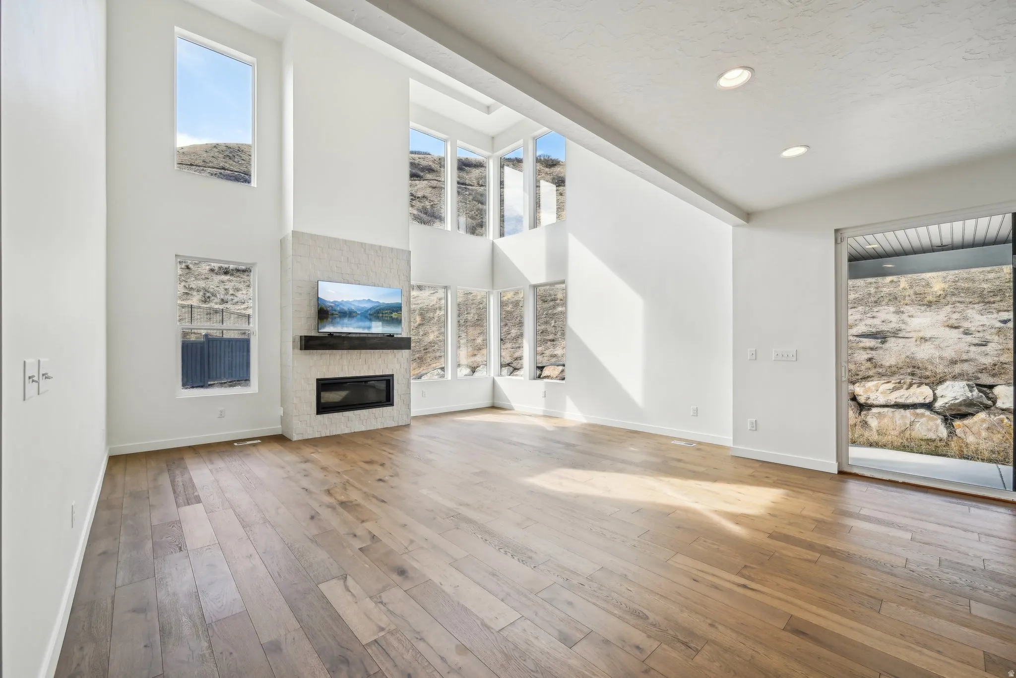 Spacious living area featuring high ceilings, wood-finish flooring, a modern fireplace with a stone-clad surround and wood mantel, recessed lighting, and numerous large windows