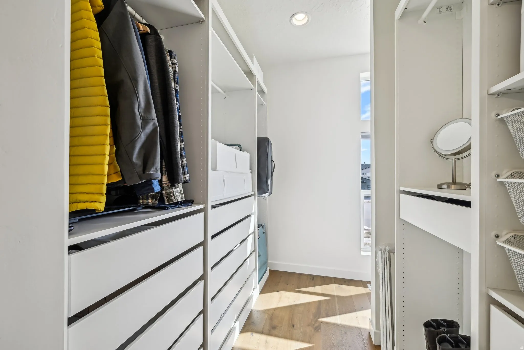 Custom closet system featuring multiple hanging sections, integrated drawer storage, open shelving, wood-finish flooring, and natural light from a vertical window