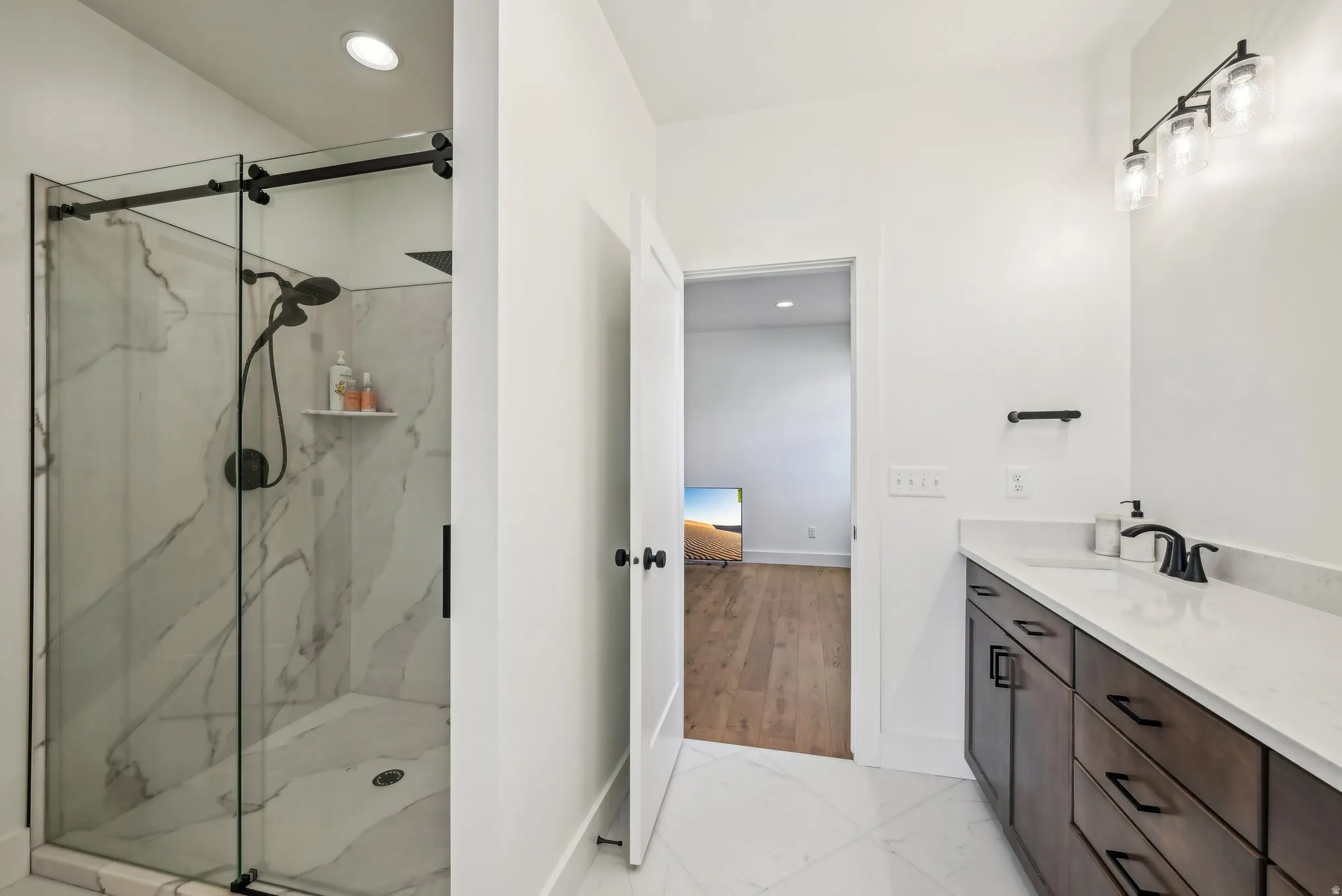 Contemporary bathroom featuring a spacious glass-enclosed shower with white marble-patterned tile, a dark wood-finish vanity with a white countertop and undermount sink, and matte black fixtures