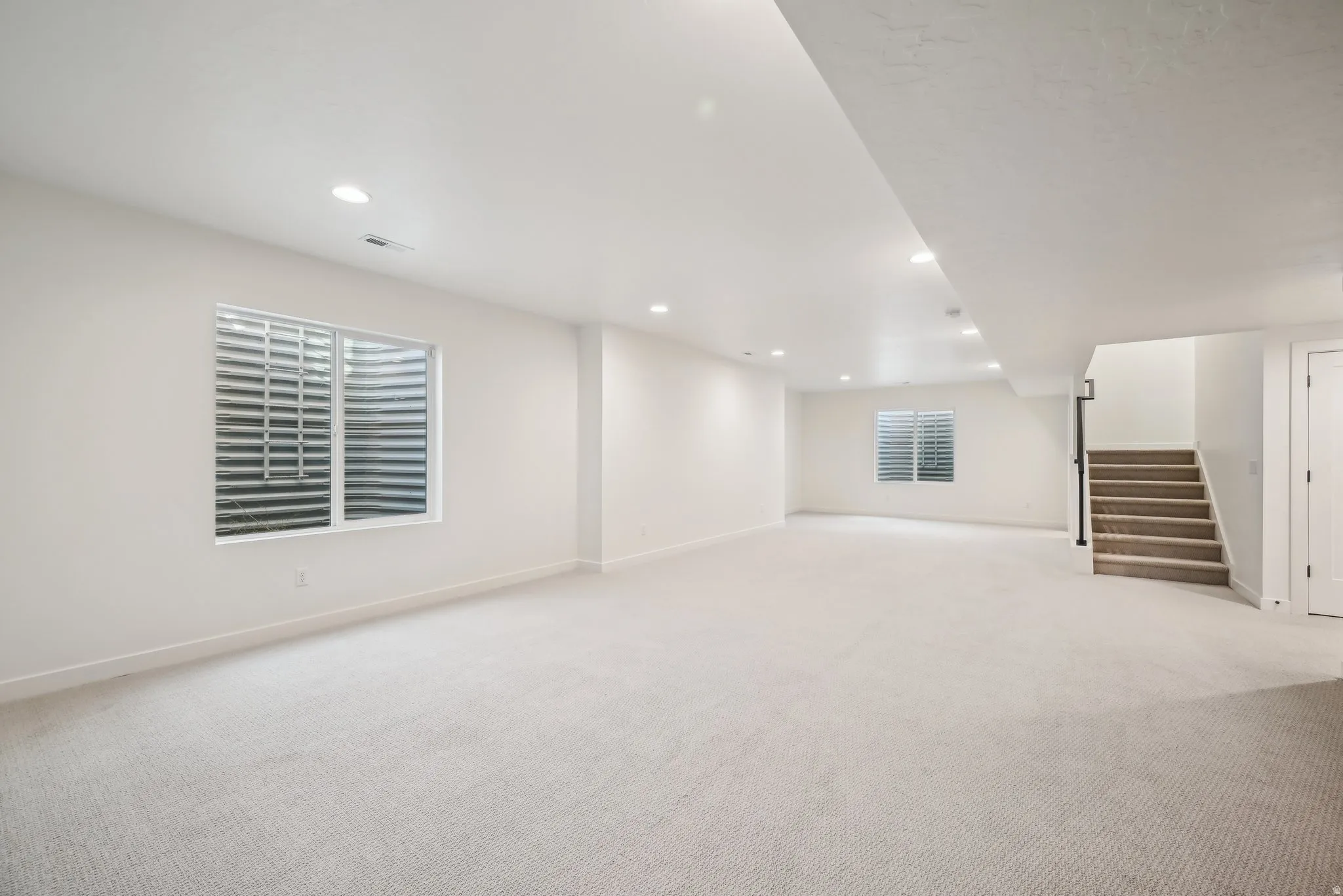 Spacious carpeted room featuring recessed lighting, two windows with horizontal blinds, and a staircase with a black metal handrail