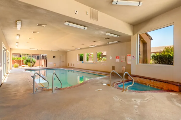 Indoor swimming pool area with an integrated spa, concrete decking, multiple windows, and fluorescent lighting fixtures