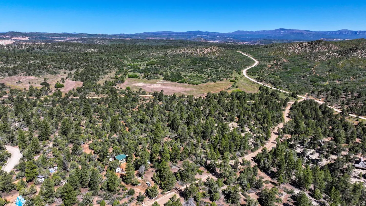 Expansive treed landscape featuring a winding unpaved road, scattered residential structures, and distant mountainous terrain under a clear sky