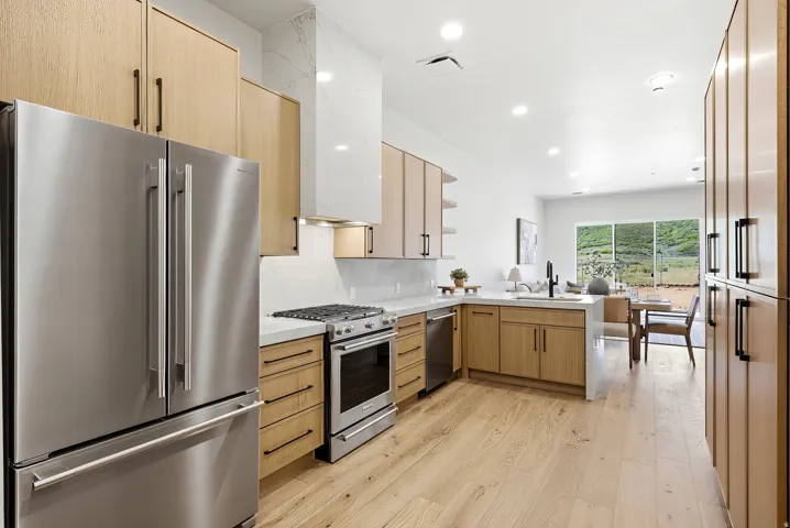 Open-concept kitchen featuring light wood-finish cabinetry, stainless steel appliances, a range hood with a white stone accent, and a light wood-finish flooring
