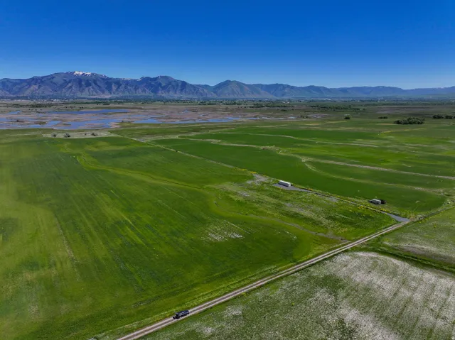Expansive green fields with a visible dirt road, featuring distant mountains with snow-capped peaks and visible bodies of water