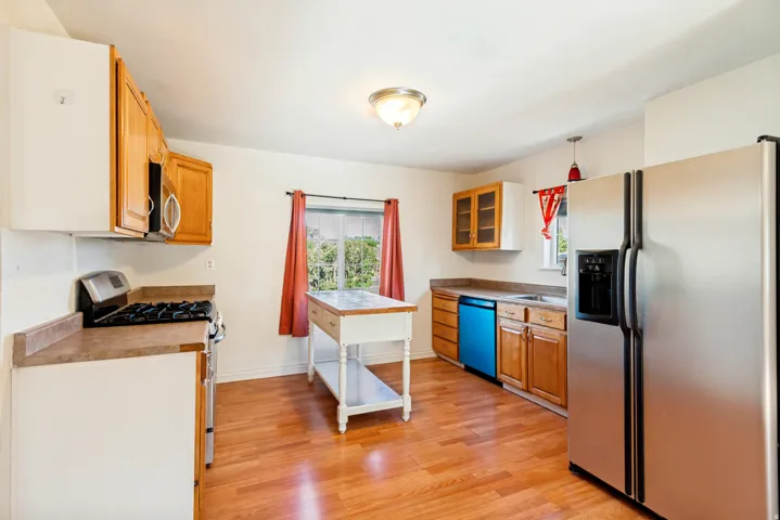 Open-concept kitchen featuring wood-finish flooring, light wood cabinetry, white perimeter cabinetry, stainless steel appliances, and a central movable island