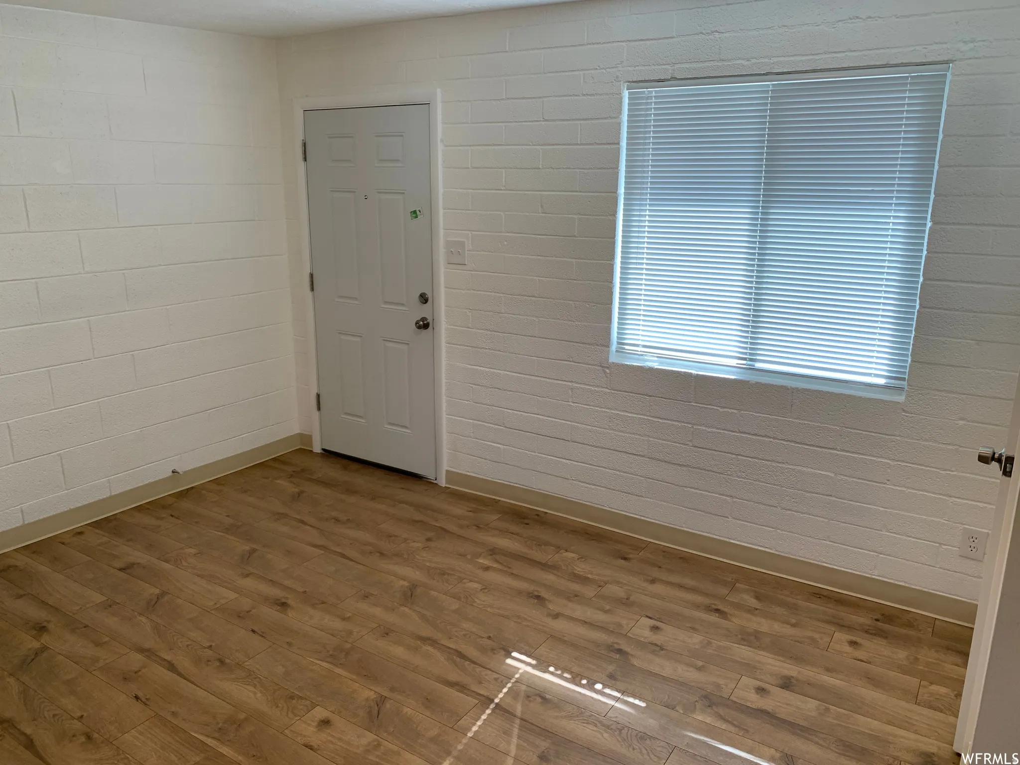Interior room featuring wood-finish flooring, white painted block walls, a white paneled door, and a window with horizontal blinds