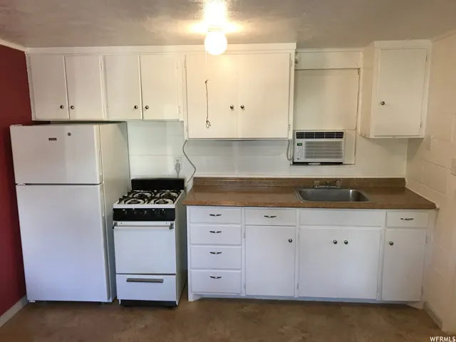 Compact kitchen featuring white cabinetry, a built-in window air conditioning unit, a white refrigerator, a white freestanding range, and a stainless steel sink with a brown countertop