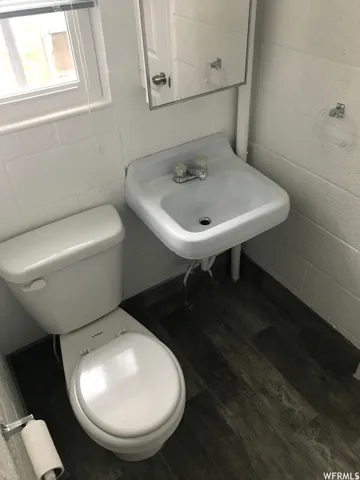 Bathroom featuring a wall-mounted sink, toilet, wood-finish flooring, and a window with blinds