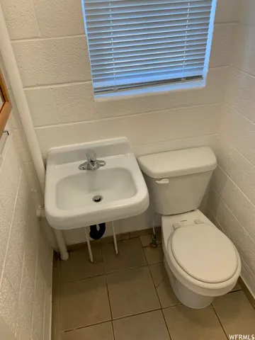 Bathroom featuring a white wall-mounted sink, a two-piece toilet, ceramic tile flooring, textured block walls, and a window with horizontal blinds