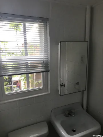 Bathroom featuring a window with horizontal blinds, a wall-mounted medicine cabinet with a mirrored door, a pedestal sink with dual faucets, and a toilet