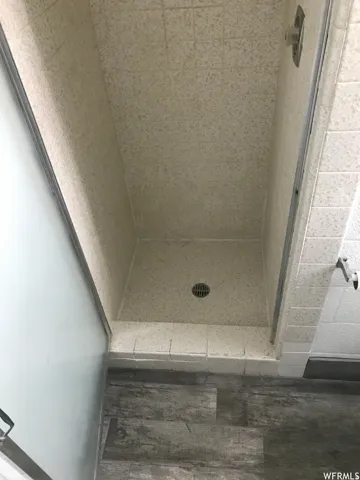 Enclosed shower stall featuring speckled beige tile walls and floor, a central drain, a chrome shower head, and wood-finish flooring outside the shower base