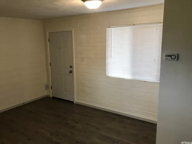 Living area featuring wood-finish flooring, painted brick walls, a window with horizontal blinds, a white paneled entry door, and a ceiling-mounted light fixture