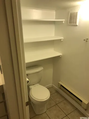 Compact bathroom featuring a white toilet, tile flooring, built-in shelving, and baseboard heating