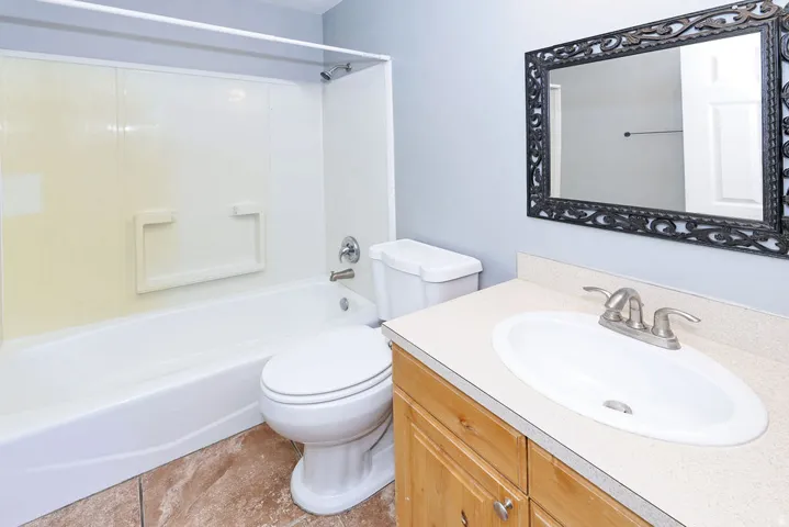 Full bathroom featuring a white bathtub/shower combination, a vanity with a light-colored countertop and an integrated sink, a wood-finish cabinet, and a decorative framed mirror