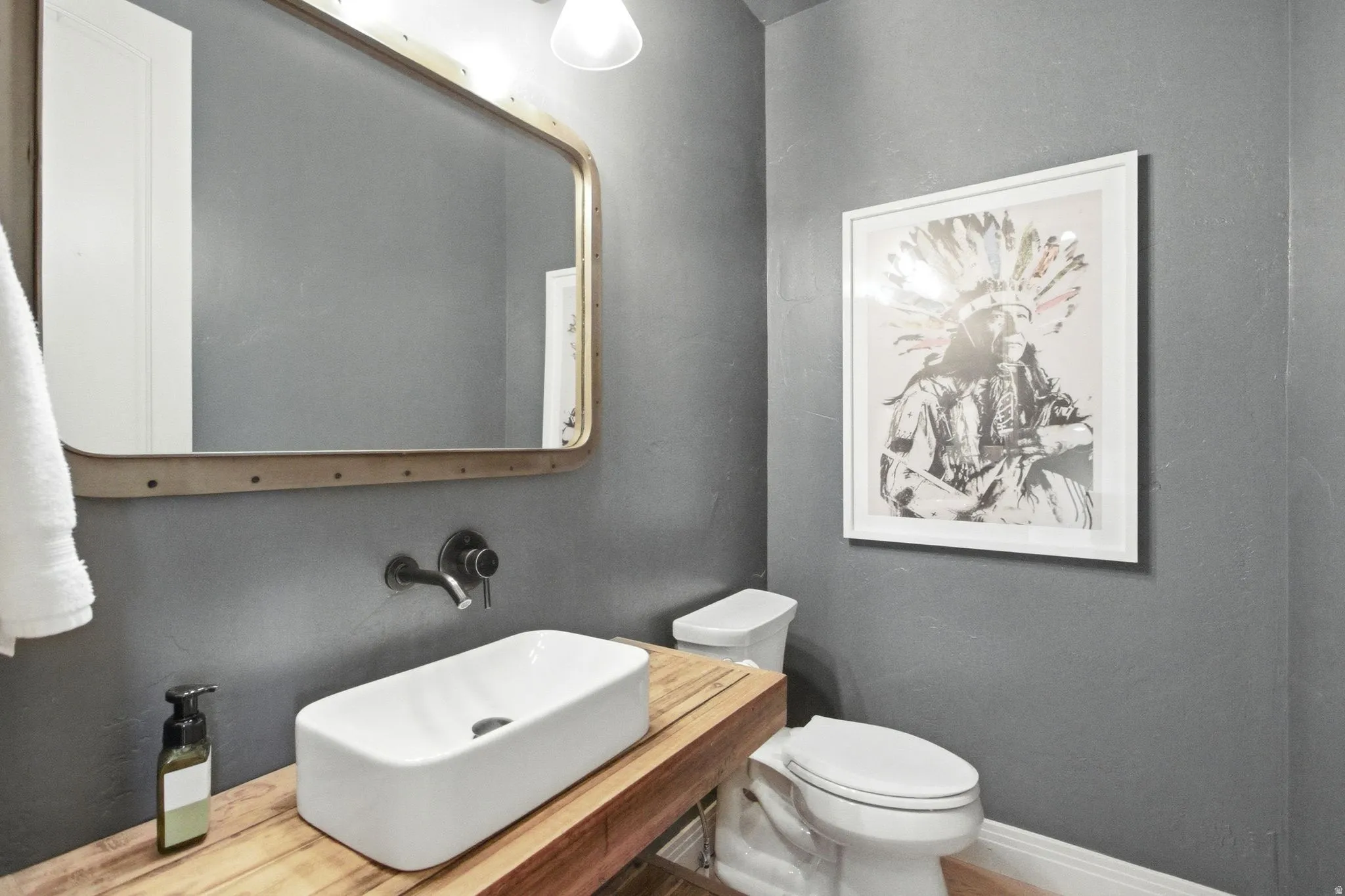 Contemporary bathroom featuring a vessel sink, wall-mounted faucet, wood countertop, industrial-style mirror, and a white toilet