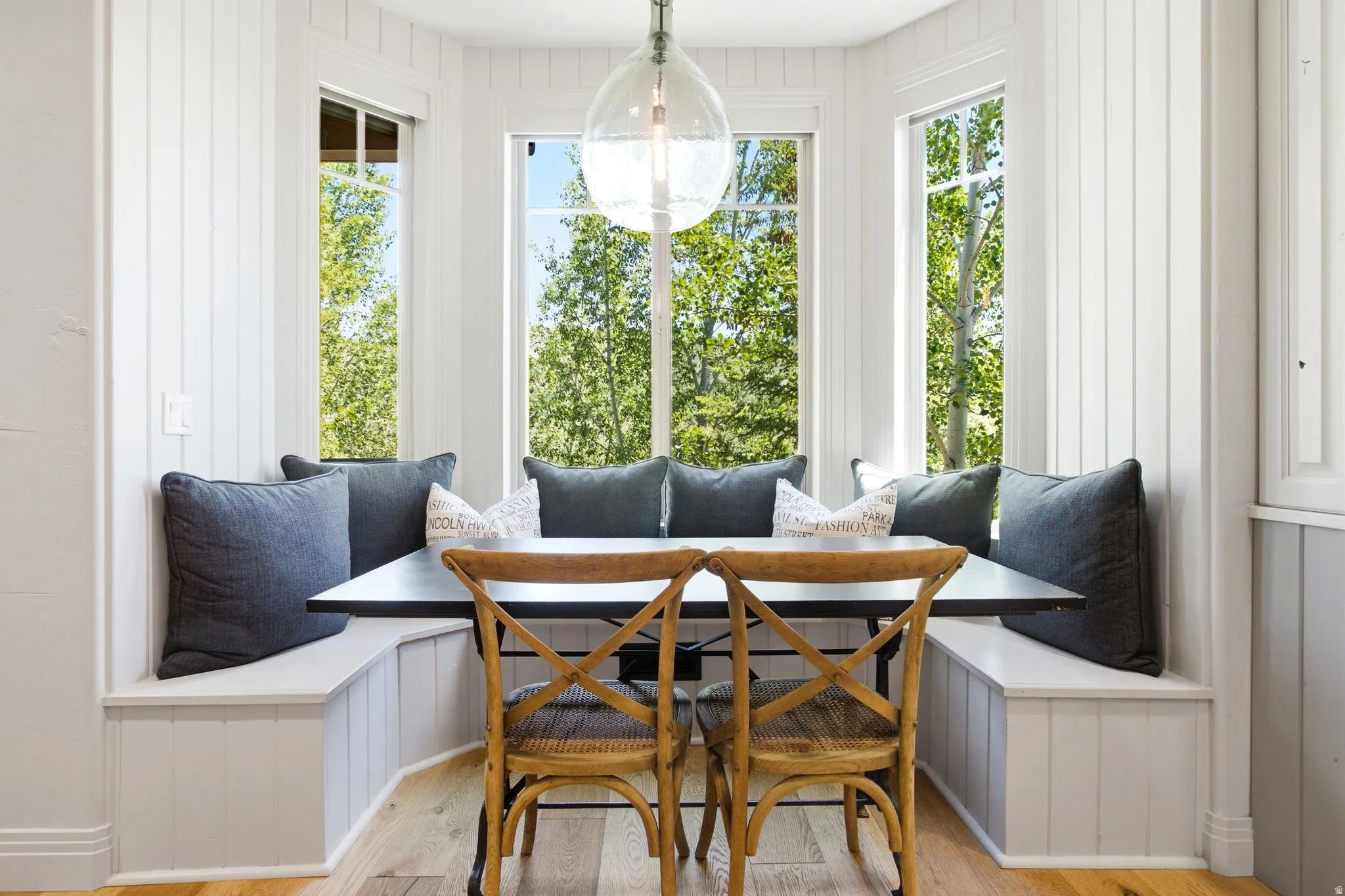 Built-in corner bench seating with white paneling, multiple large windows, a black table with a dark metal base, an overhead glass pendant light fixture, and wood-finish flooring