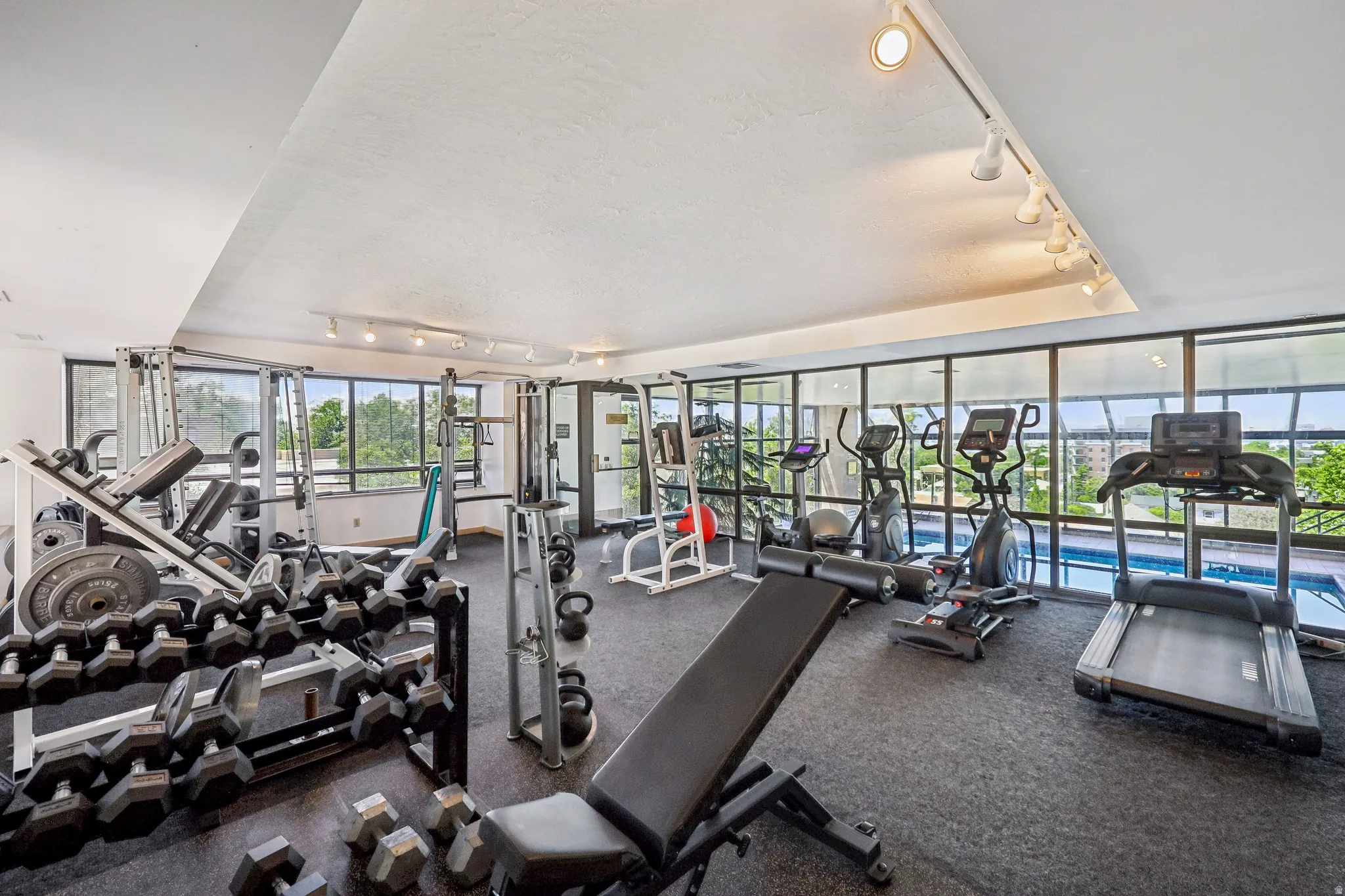 Fitness center with track lighting, expansive windows, cushioned flooring, and an adjacent indoor pool