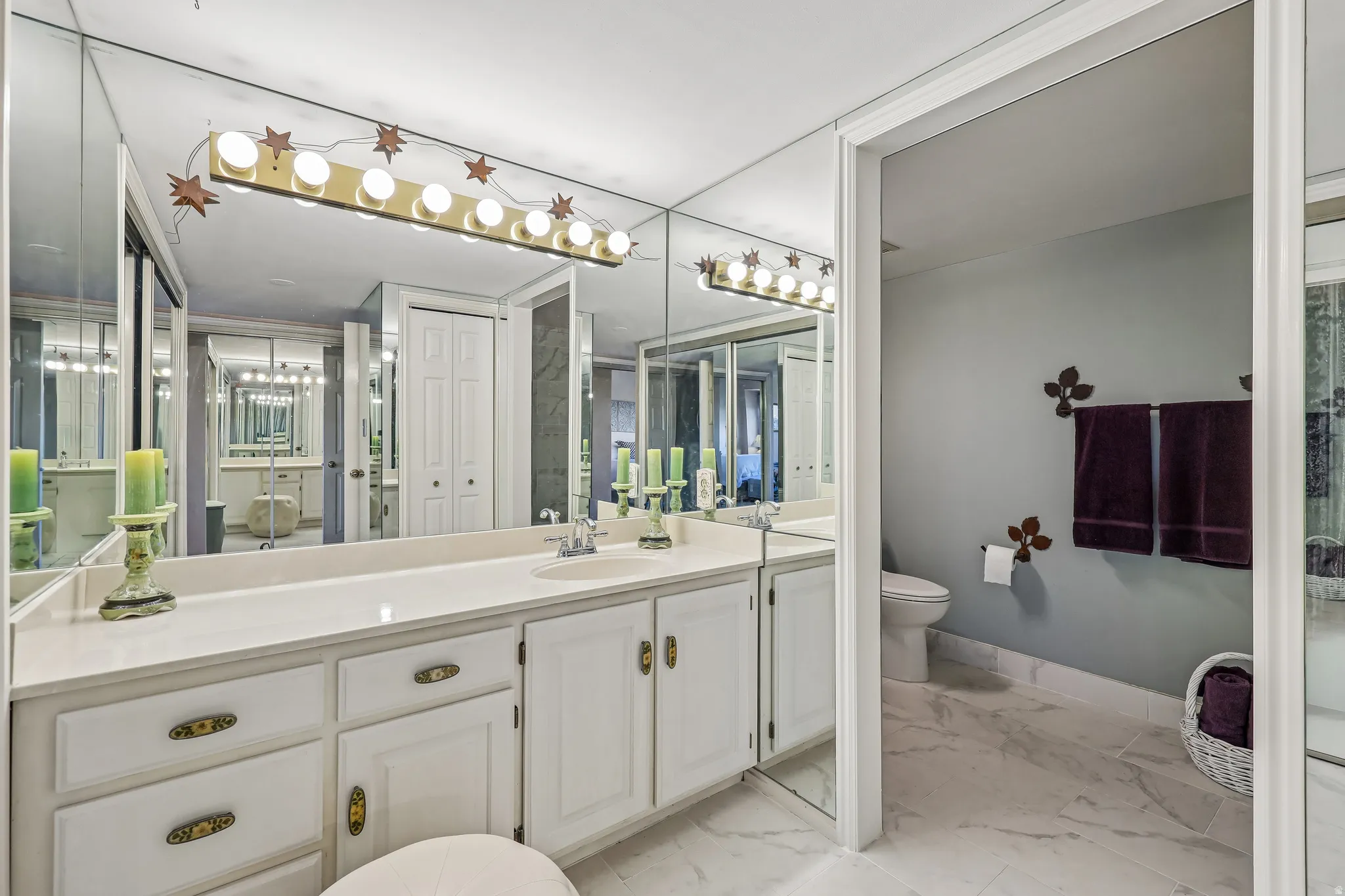 Bathroom vanity featuring extensive mirrored walls, a wide countertop, an integrated sink, white cabinetry with decorative hardware, and a multi-bulb lighting fixture