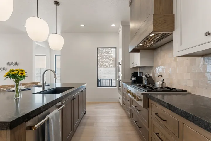 Contemporary kitchen featuring a spacious island with an undermount sink, pendant lighting, professional-grade range, custom wood-finish cabinetry, and light-toned tile backsplash