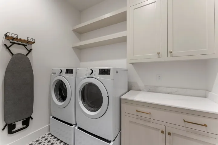 Laundry area featuring patterned floor tiles, light-toned shaker-style cabinetry with gold-tone hardware, white countertop, and open shelving
