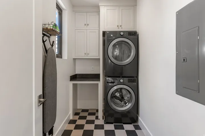 Laundry area featuring stacked appliances, black and white checkered flooring, a built-in counter with dark stone-finish surface, and white upper cabinetry