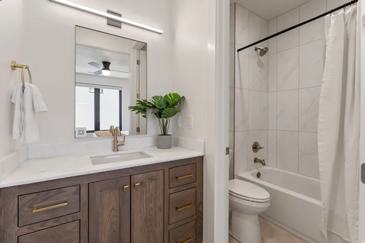 Bathroom featuring a wood-finish vanity with gold-tone hardware, a white countertop with an integrated rectangular sink, a wall-mounted rectangular mirror, and a linear overhead light fixture